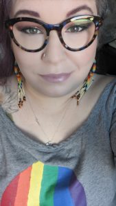 woman with purple hair and colorful glasses wearing rainbow dreamcatcher earrings and a gray shirt with a rainbow skull